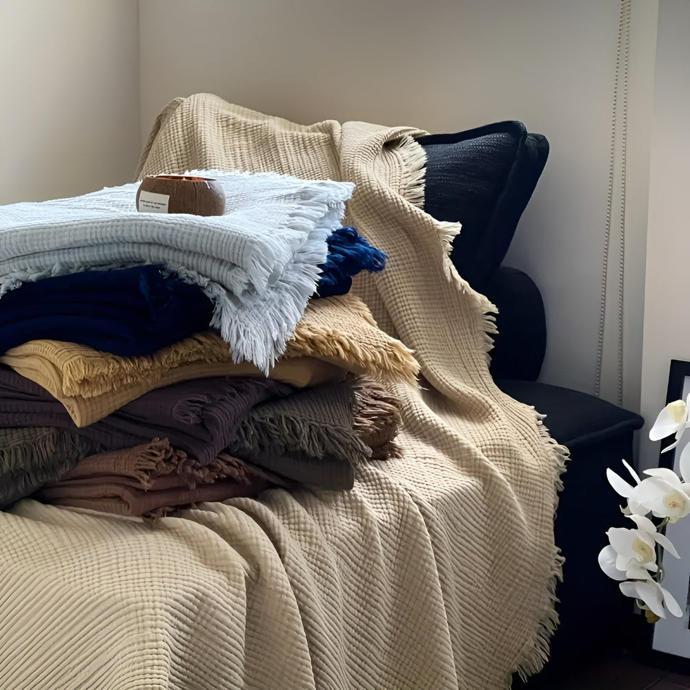 Stack of crinkle cotton blankets in various colors, including sand, navy, and light blue, with tassels.