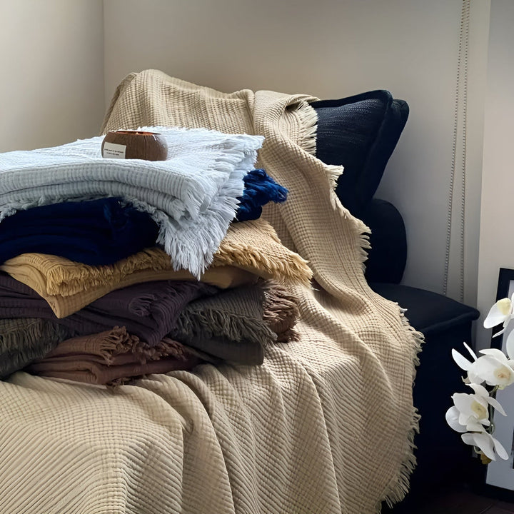Stack of crinkle cotton blankets in various colors, including sand, navy, and light blue, with tassels.