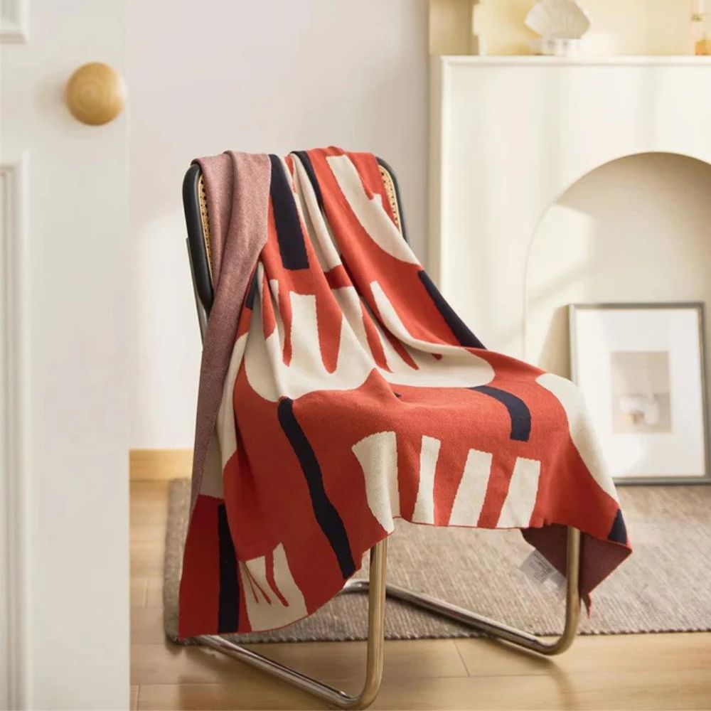 A chair draped with a red, cream, and navy floral knitted blanket and a pink throw.
