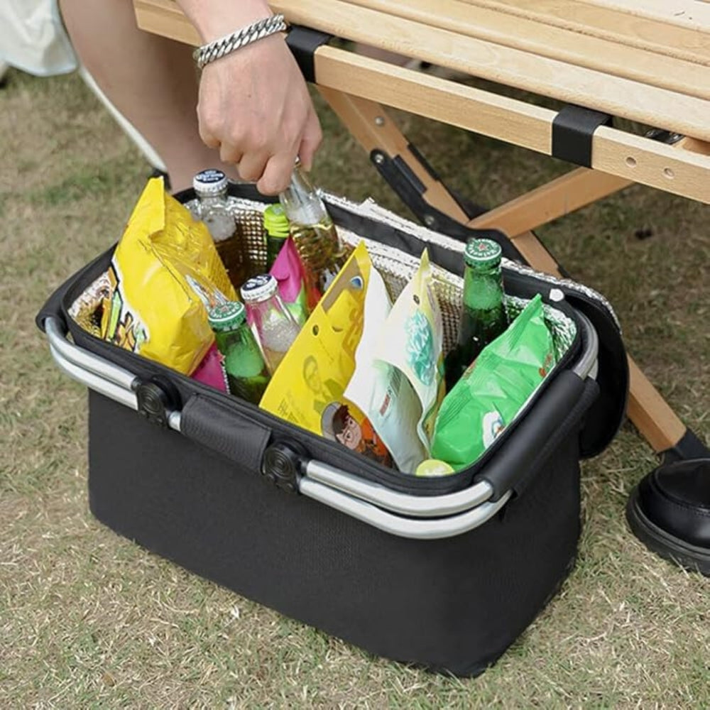 Black folding picnic basket filled with snacks and bottles, placed on grass next to a wooden table.