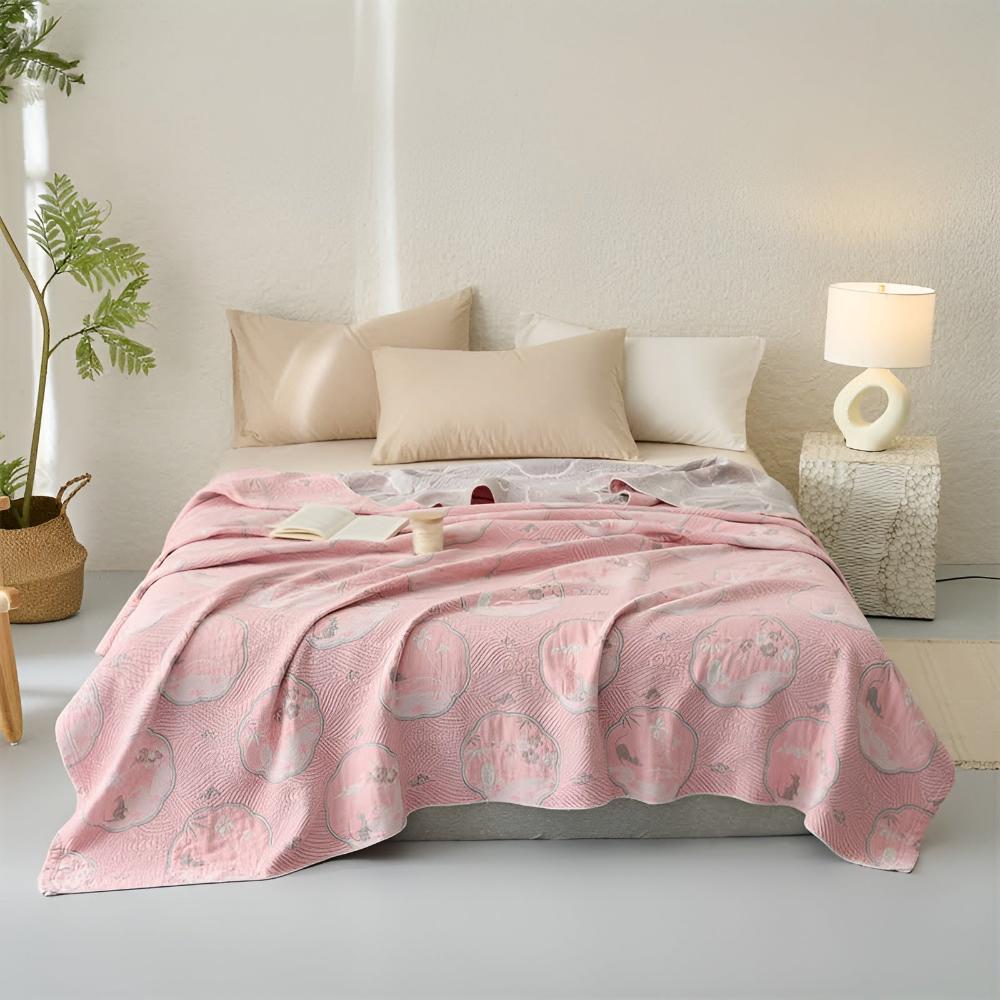 Pink reversible blanket with a circular pattern draped over a bed with pillows and a lamp.