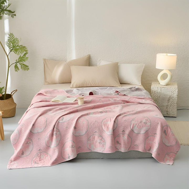 Pink reversible blanket with a circular pattern draped over a bed with pillows and a lamp.