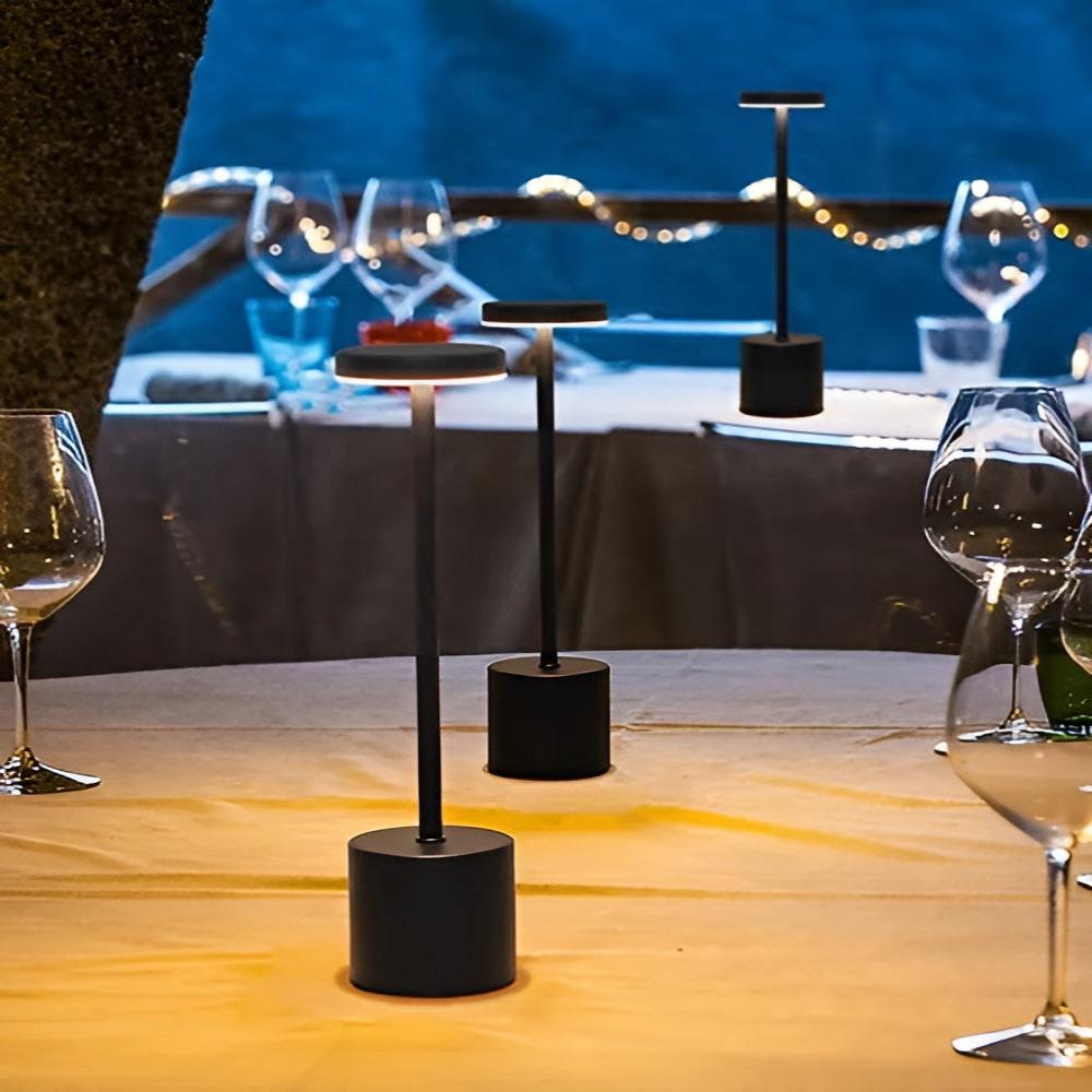 Three black cordless table lamps with warm light on a table set for dinner outdoors.