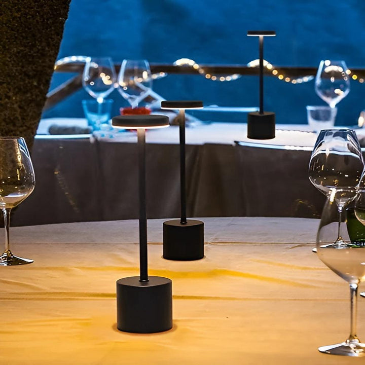 Three black cordless table lamps with warm light on a table set for dinner outdoors.