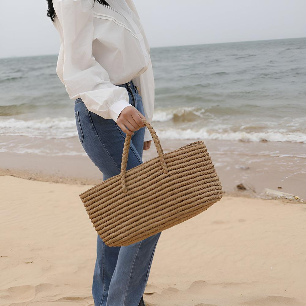 Woman holding a handwoven straw tote bag on a sandy beach with the ocean in the background.