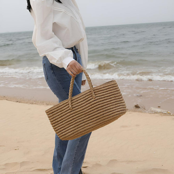 Woman holding a handwoven straw tote bag on a sandy beach with the ocean in the background.