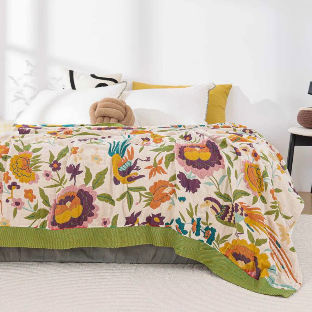 A lightweight coverlet with a vibrant floral and bird pattern in shades of yellow, orange, purple, and green on a cream background.