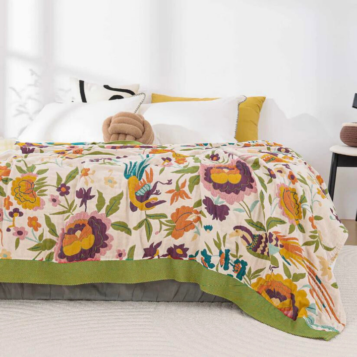 A lightweight coverlet with a vibrant floral and bird pattern in shades of yellow, orange, purple, and green on a cream background.