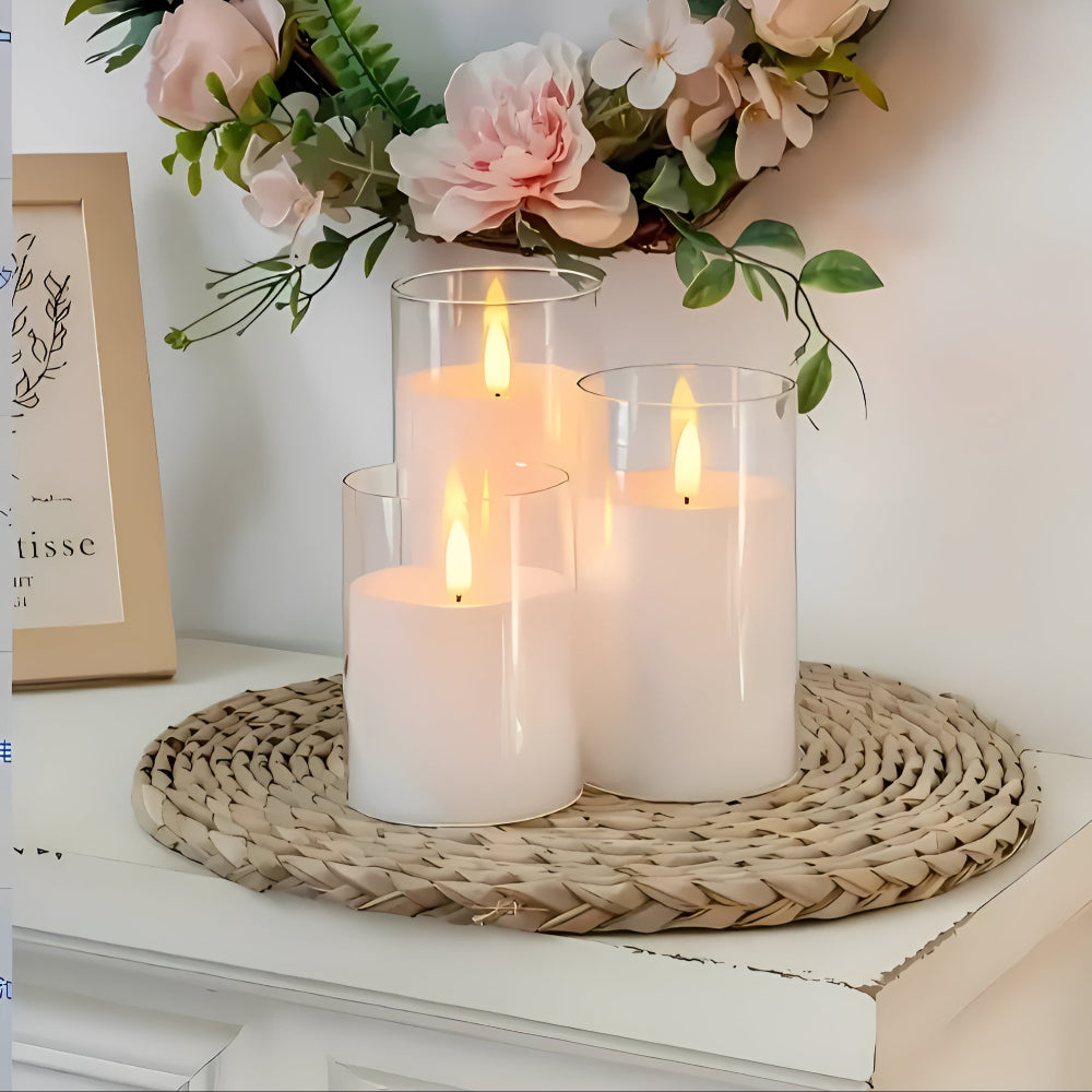 Three white flameless LED candles of varying heights in glass cylinders on a woven mat.