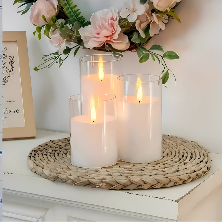 Three white flameless LED candles of varying heights in glass cylinders on a woven mat.