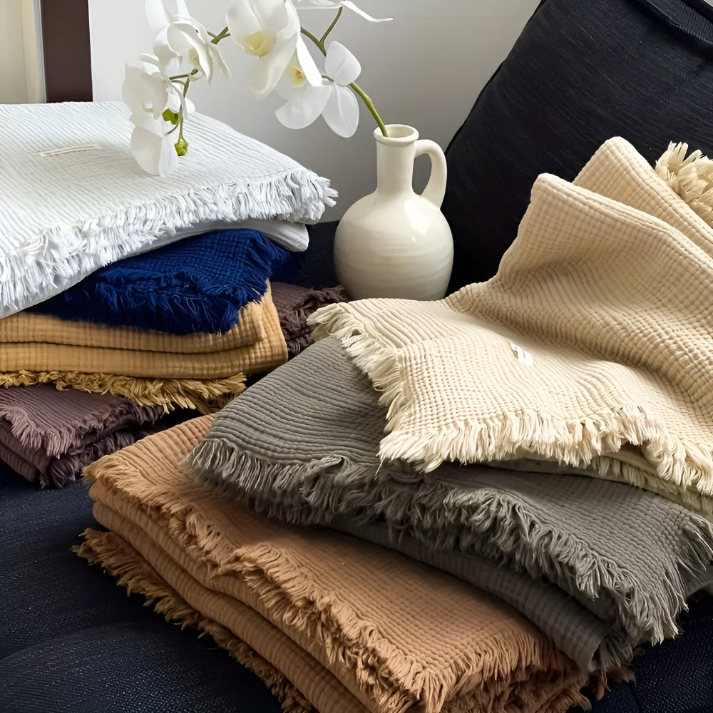Stacks of cotton crinkle tassel blankets in various colors: white, blue, tan, grey, and cream.