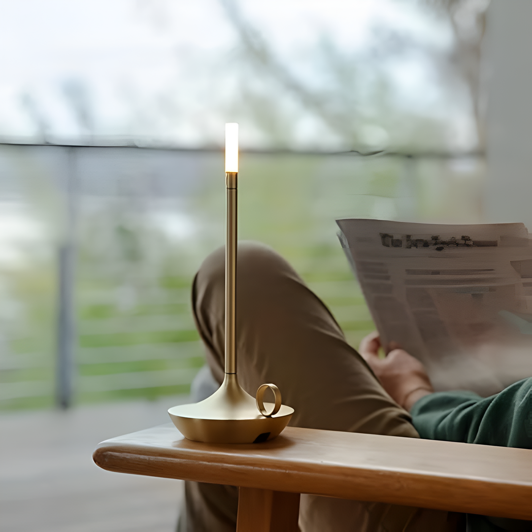 Portable aluminum table lamp with a warm glow, placed on a wooden table next to a person reading a newspaper.