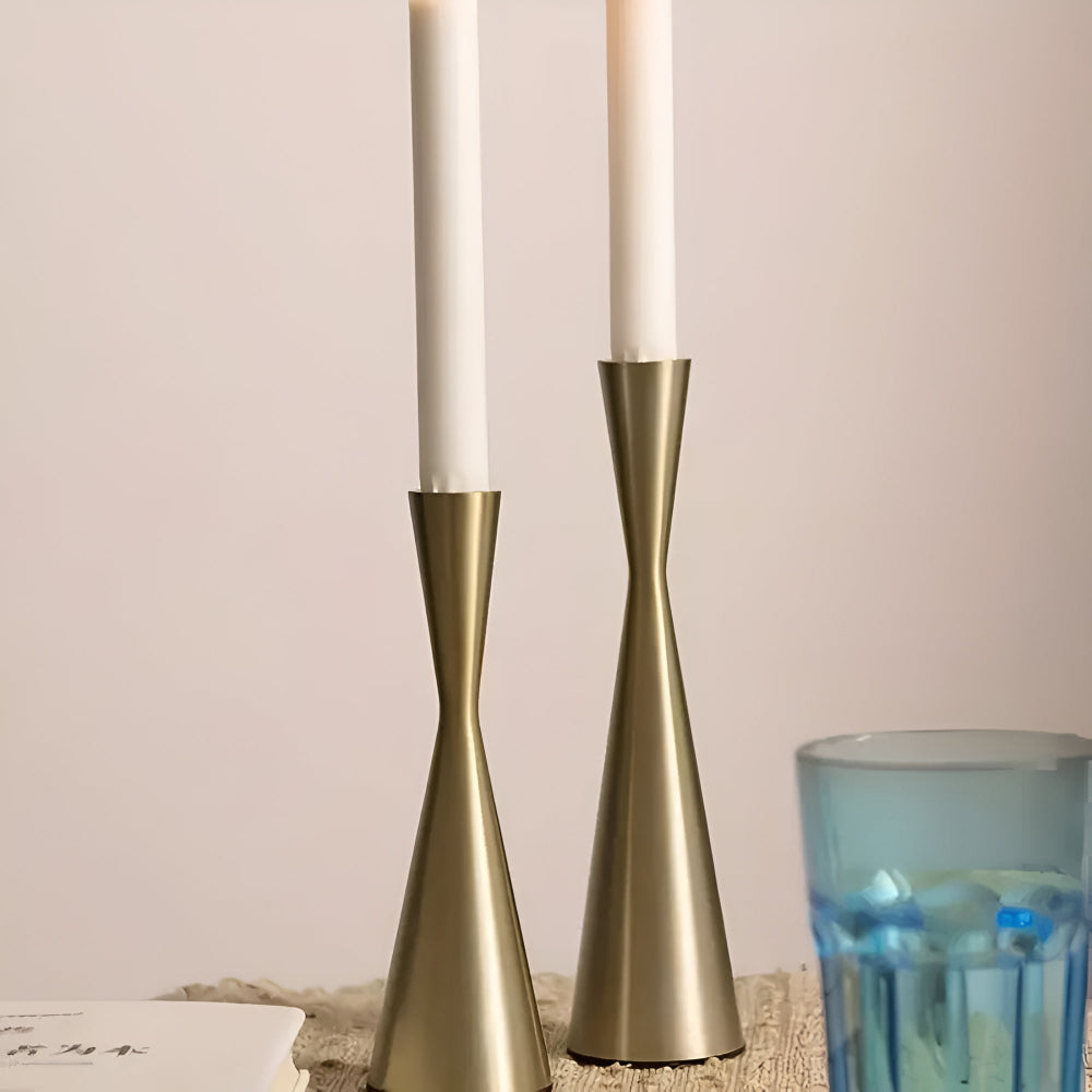Two brass candlesticks with white candles on a textured surface, with a blue glass in the foreground.