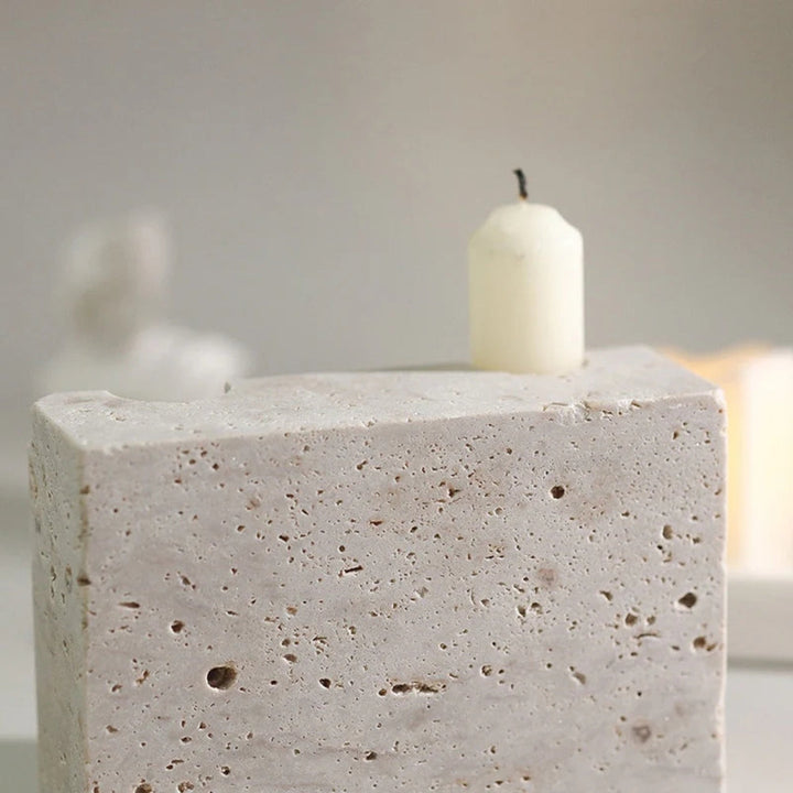 A short, cream-colored candle sits atop a beige travertine stone block with visible pores.