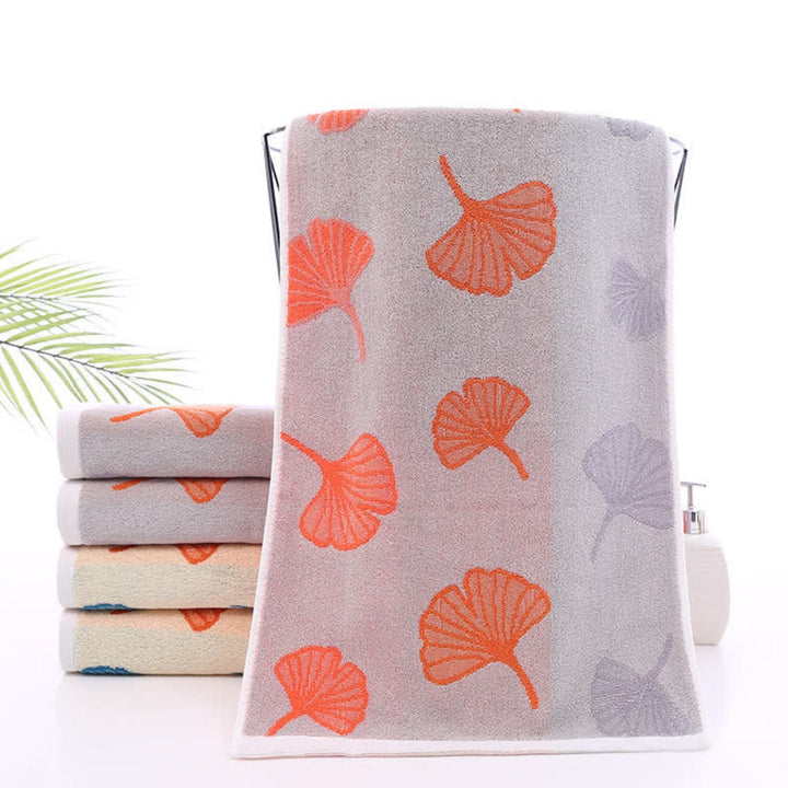 Grey bath towel with orange and purple ginkgo leaf pattern, stacked towels in background.