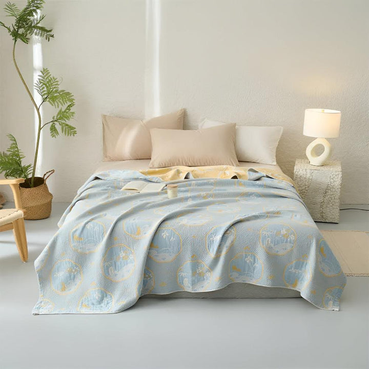 Reversible light blue blanket with a coastal pattern draped over a bed with pillows and a lamp.