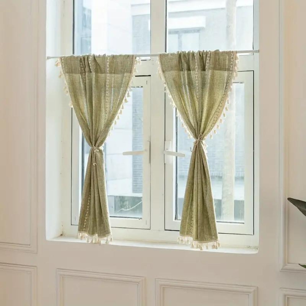 Two green boho cotton linen kitchen curtains with white tassels hanging on a window.