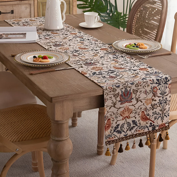 Vintage floral and bird jacquard table runner with tassels on a wooden table.