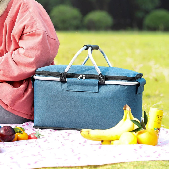 Blue folding picnic basket with handles, filled with fruit and a can of drink, on a picnic blanket.