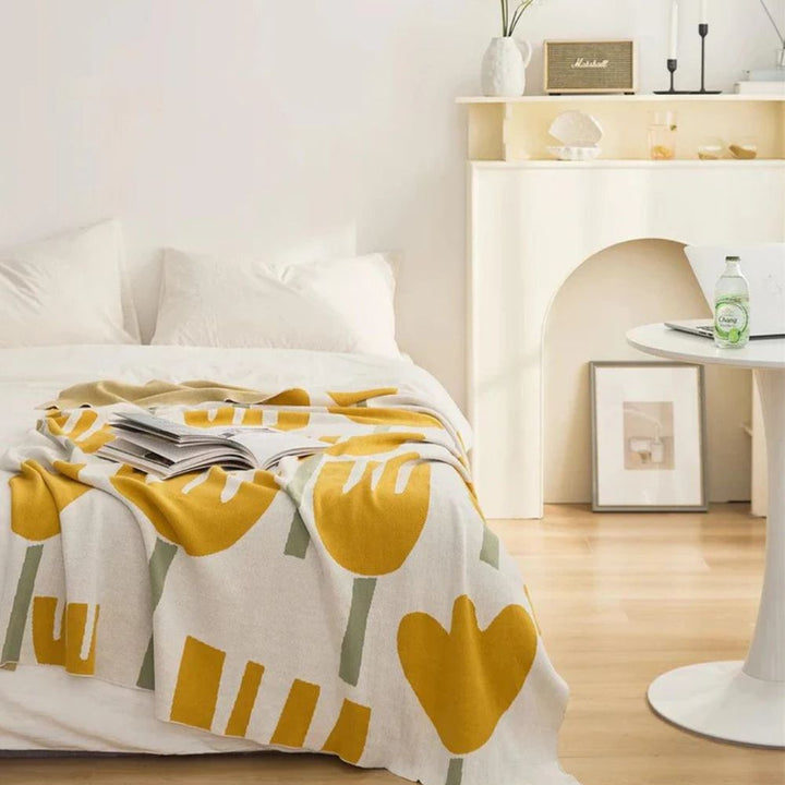 A knitted blanket with a yellow tulip floral pattern draped over a white bed.