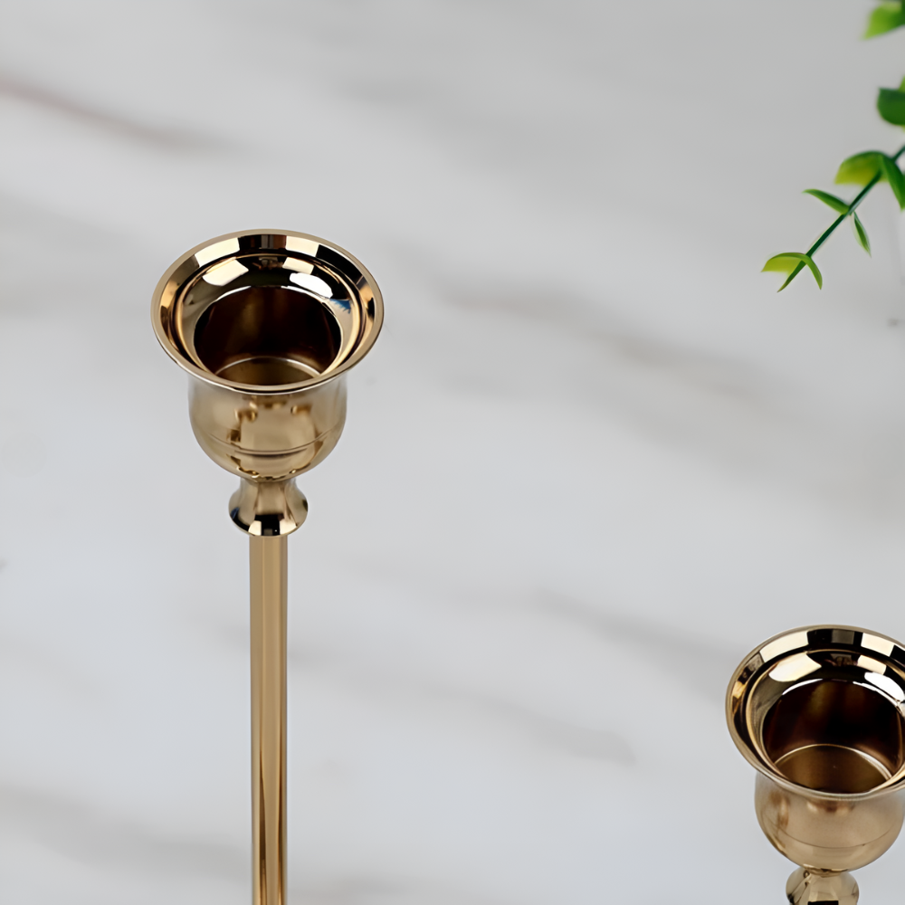 Two gold taper candle holders on a white marble surface with a green plant in the background.