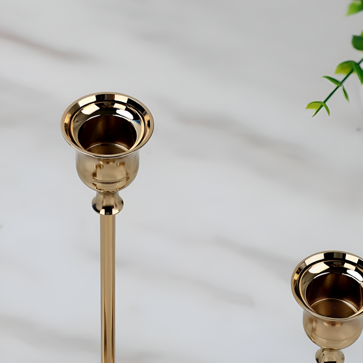 Two gold taper candle holders on a white marble surface with a green plant in the background.
