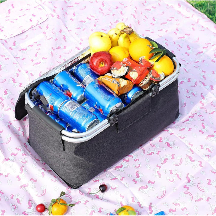 Folding picnic basket filled with cans of soda, fruit, and snacks on a unicorn-patterned blanket.
