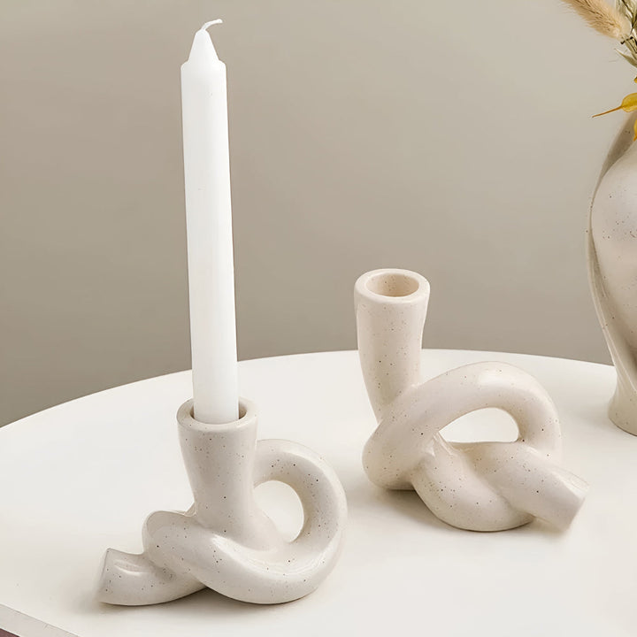 Two cream-colored ceramic candle holders with a knotted design, one holding a white taper candle.