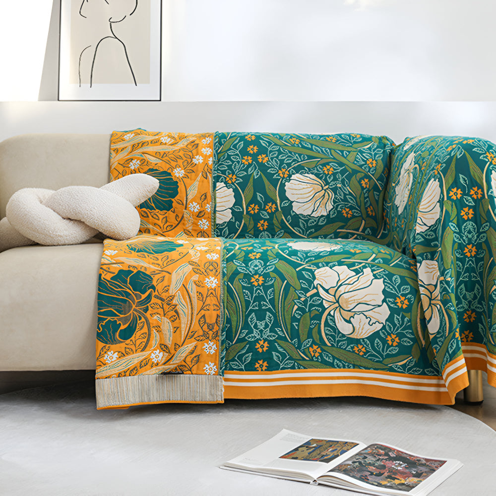 A couch draped with a green botanical bloom cotton blanket and an orange floral blanket with fringe.