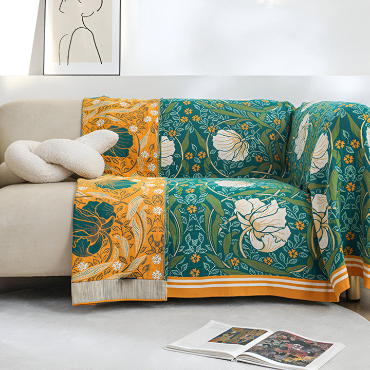 A couch draped with a green botanical bloom cotton blanket and an orange floral blanket with fringe.