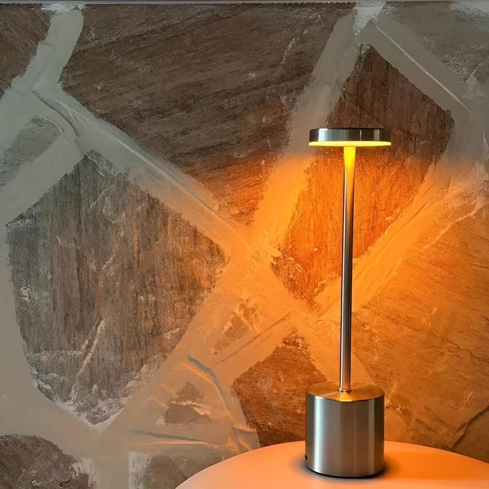 A cordless table lamp with a brushed metal base and stem, emitting a warm yellow light against a textured stone wall.