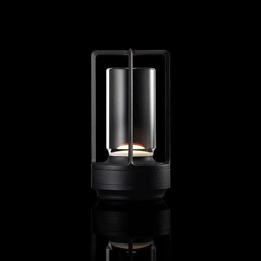 Black table lantern with a cylindrical glass shade and a metal frame, emitting a warm glow.