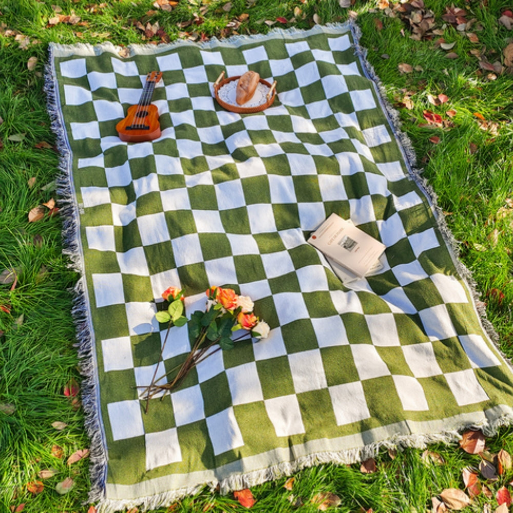 Green and white checkered picnic blanket with fringe, laid out on grass with a book, flowers, and bread.