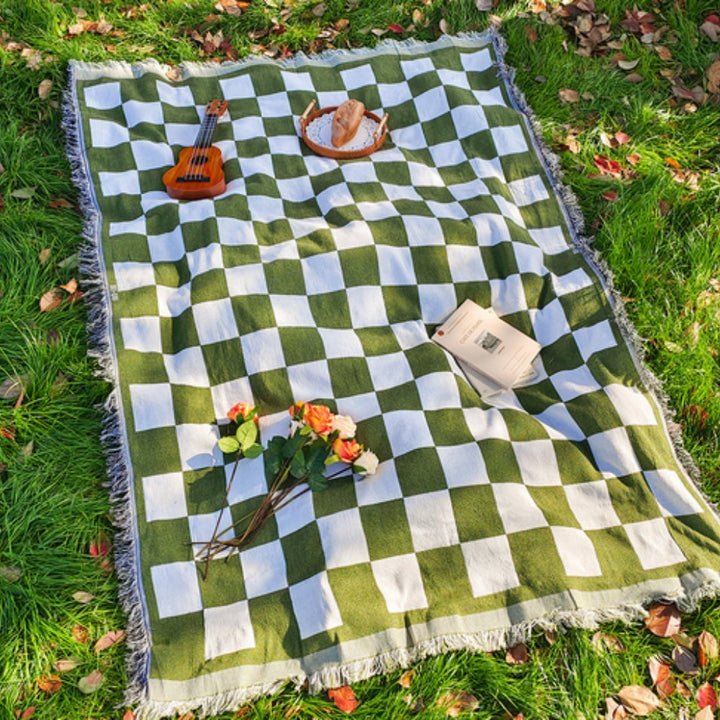 Green and white checkered picnic blanket with fringe, laid out on grass with a book, flowers, and bread.