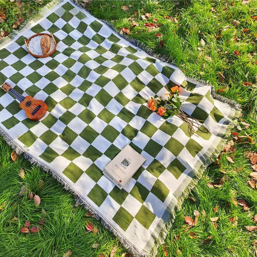 Green and white checkered picnic blanket with fringe, ukulele, bread, and flowers on grass.