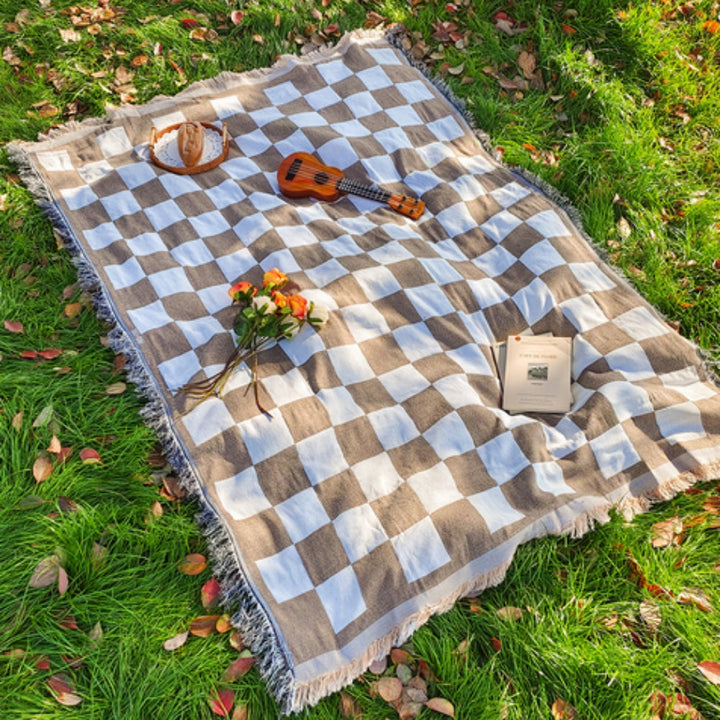 Brown and white checkered picnic blanket with fringe, laid out on grass with a ukulele, bread, flowers, and a book.