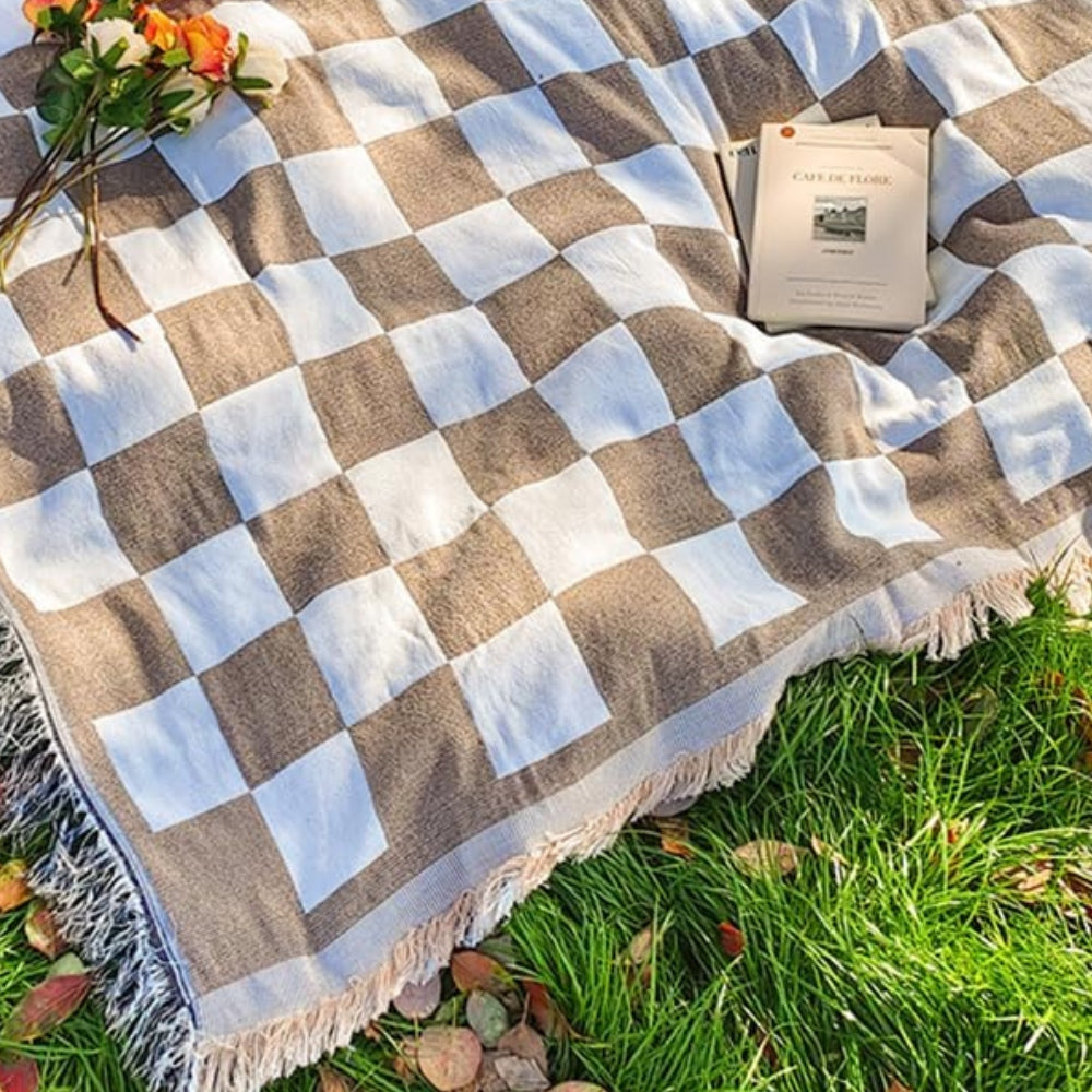 Brown and white checkered picnic blanket with fringe, laid on grass with flowers and a book.