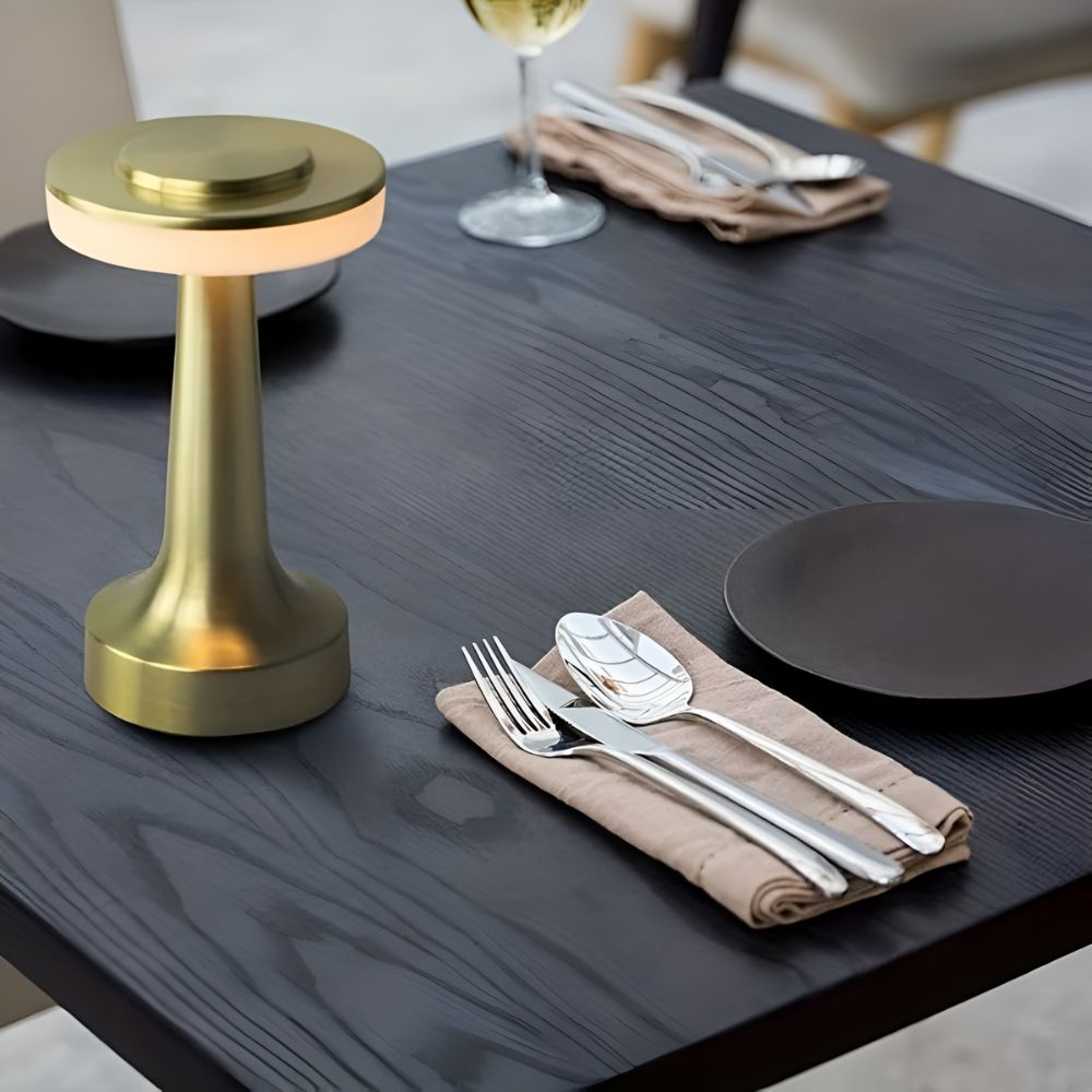 Gold cordless table lamp with a warm glow on a dark wood table with place settings.