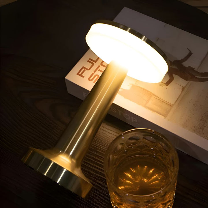 Brass cordless table lamp with a glowing dome top sits on a dark wood table next to a glass.