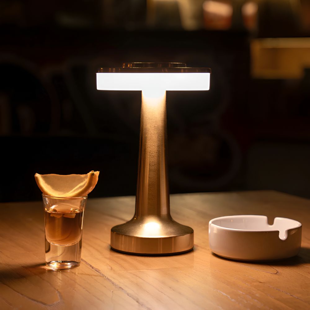 A gold cordless table lamp with a warm glow sits on a wooden table next to a shot glass and an ashtray.
