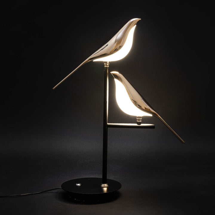 Two gold bird-shaped lamps with internal LED lights on a black stand against a black background.