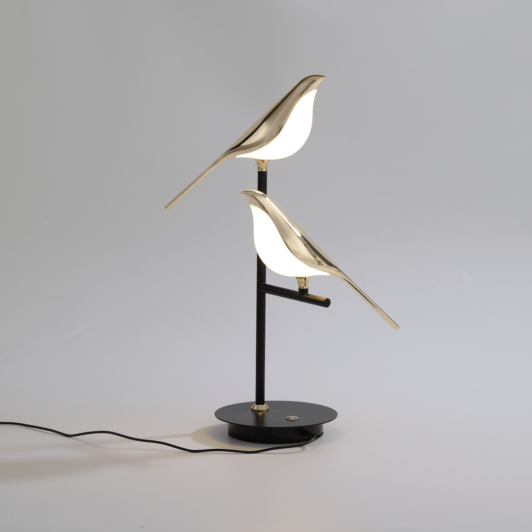 Modern Nordic table lamp with two polished metal birds perched on a black stem, emitting a warm LED glow.