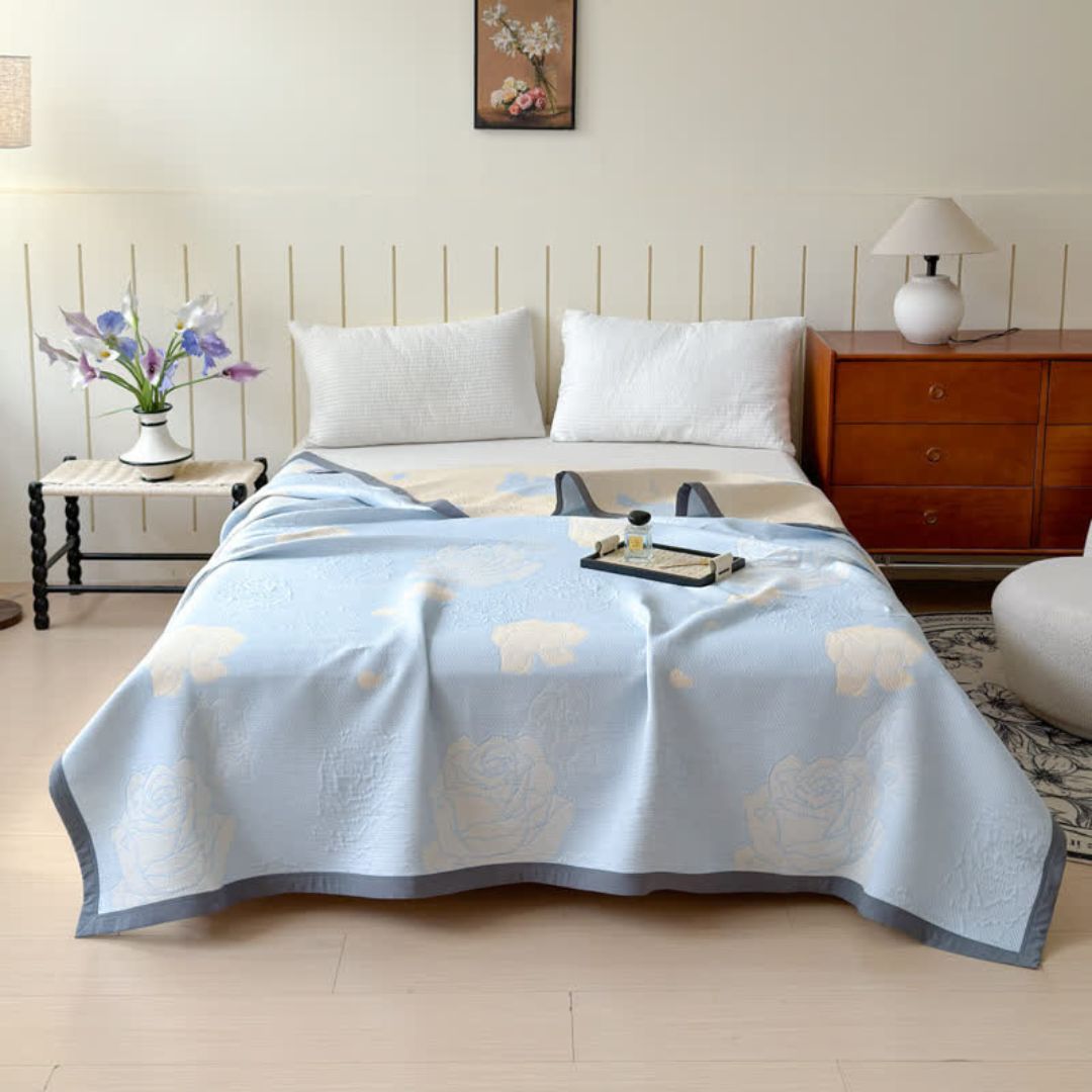 Blue rose bloom cotton blanket draped on a bed with white pillows and a bedside table.