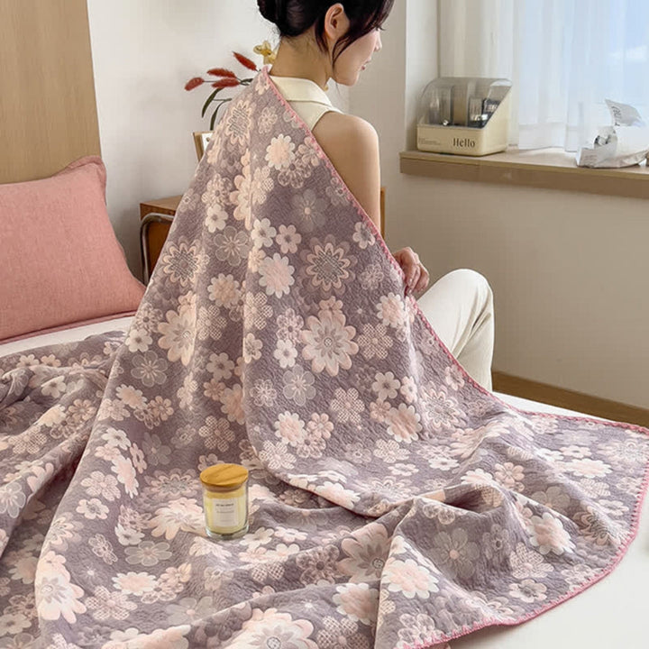 A woman sits on a bed covered by a floral cotton gauze blanket with pink and gray flowers.