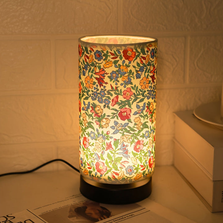 Cylindrical table lamp with a botanical floral fabric shade and a black base, emitting warm light.