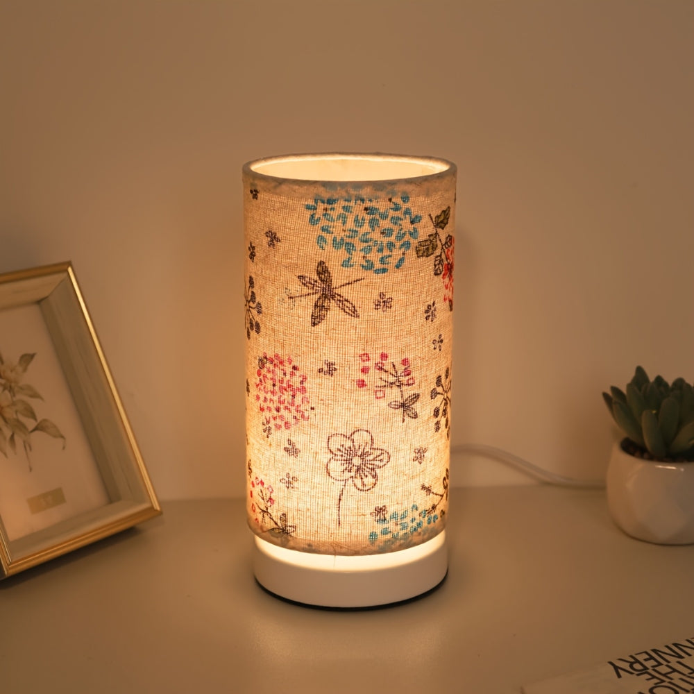 Botanical floral table lamp with a fabric shade featuring dragonflies and flowers, casting a warm glow.