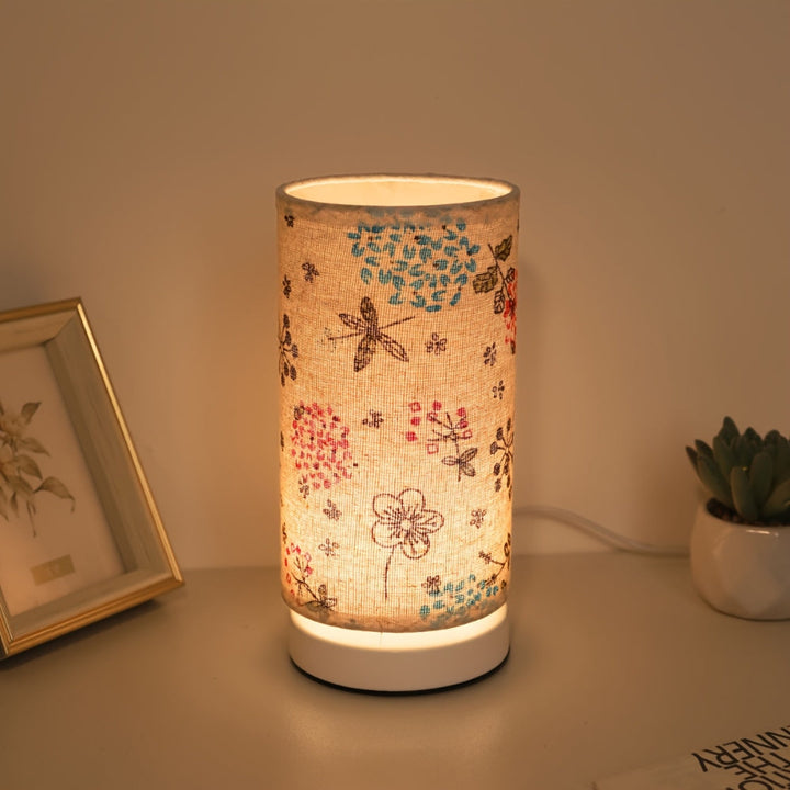 Botanical floral table lamp with a fabric shade featuring dragonflies and flowers, casting a warm glow.