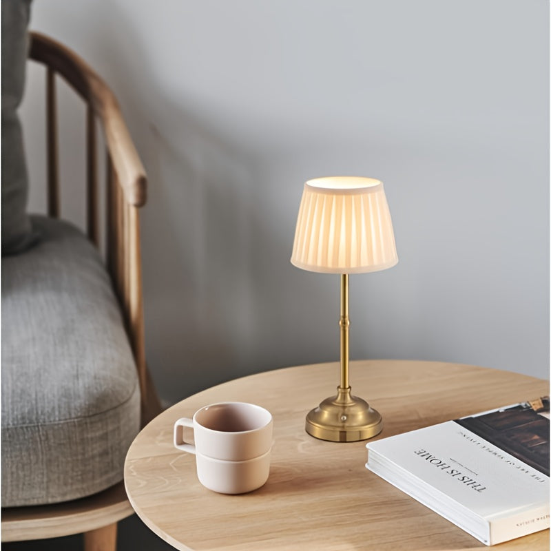 Brass cordless rechargeable table lamp with pleated shade on a wooden table next to a mug.