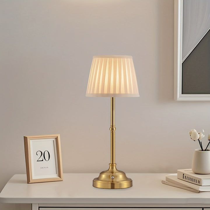Brass cordless table lamp with pleated shade on a white table.
