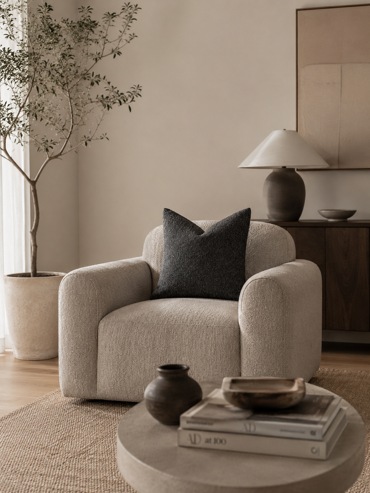 A textured neutral pillow rests on a plush armchair in a minimalist living room.
